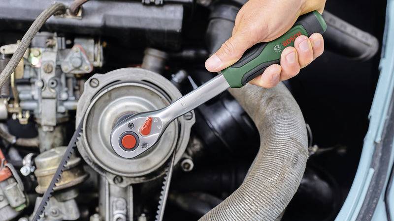 A hand is holding a green spanner and tightening a screw in the engine compartment of a car to carry out maintenance work.