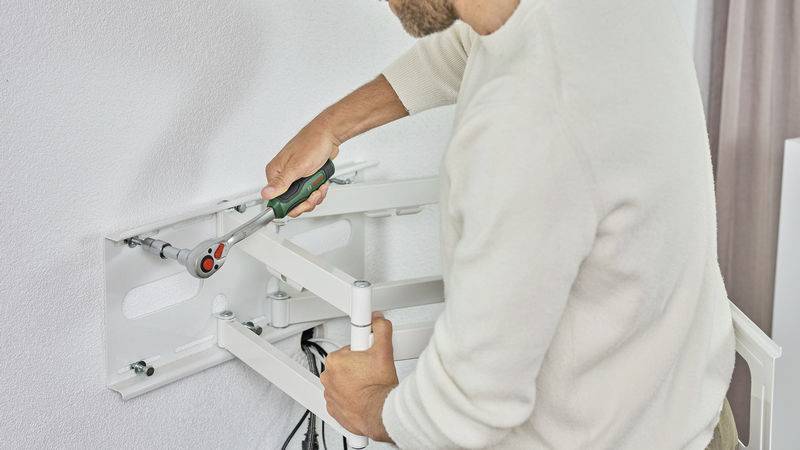 A person is mounting a white TV wall bracket on an interior wall using a screwdriver.