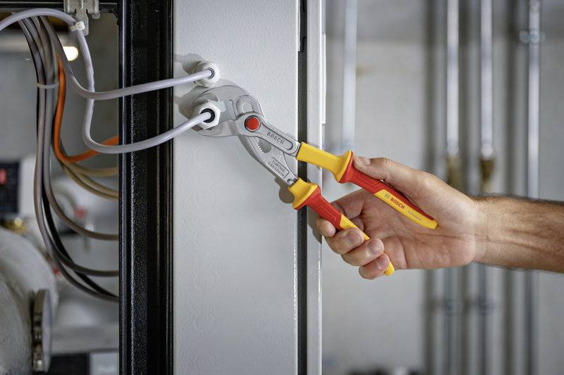 A hand holds an adjustable spanner and pulls on white cables in an electrical junction box. Further cables are visible in the background.