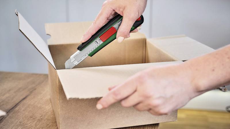 A person is opening a cardboard box with a craft knife. Hands and tool are visible.