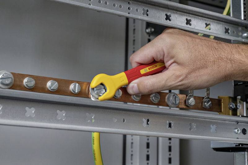 A hand holds a yellow-red spanner and tightens a nut on a metal rail inside an electrical control cabinet.