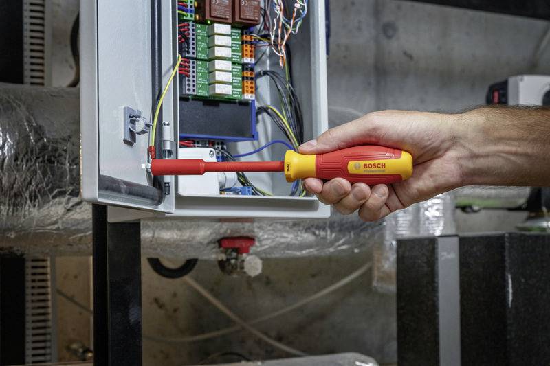 A hand holds a screwdriver working on an open electrical cabinet. Inside, colourful cables and circuit boards are visible.