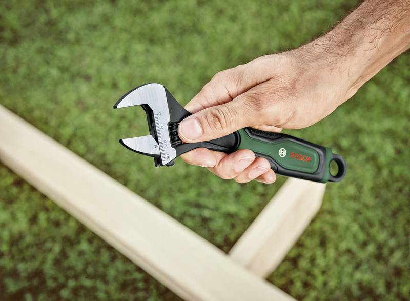 A hand holds an adjustable spanner outdoors, with a blurred wooden frame and grass in the background. A tool by Bosch.