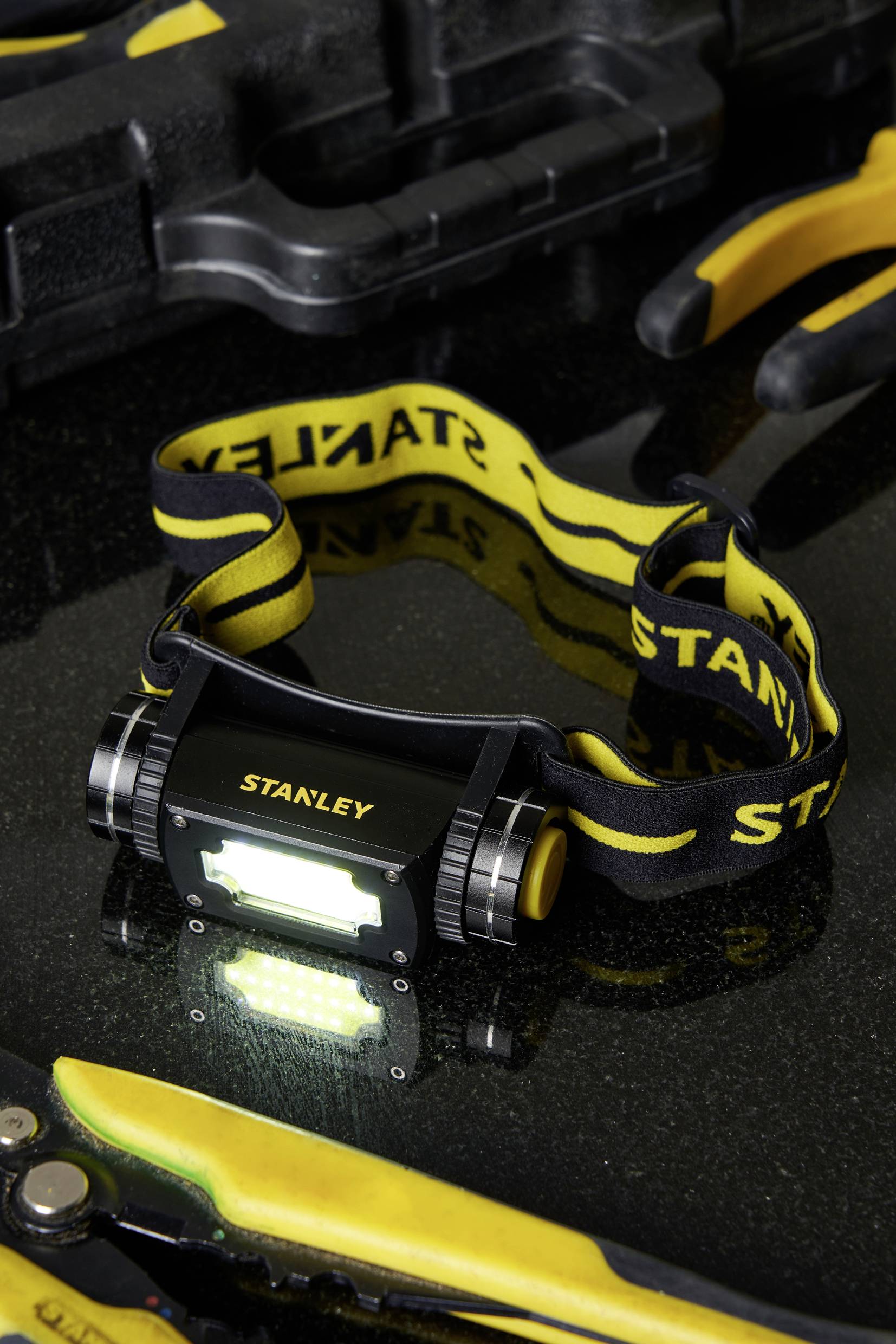 Head torch with yellow and black band rests on a black surface, surrounded by tools. Light is switched on.