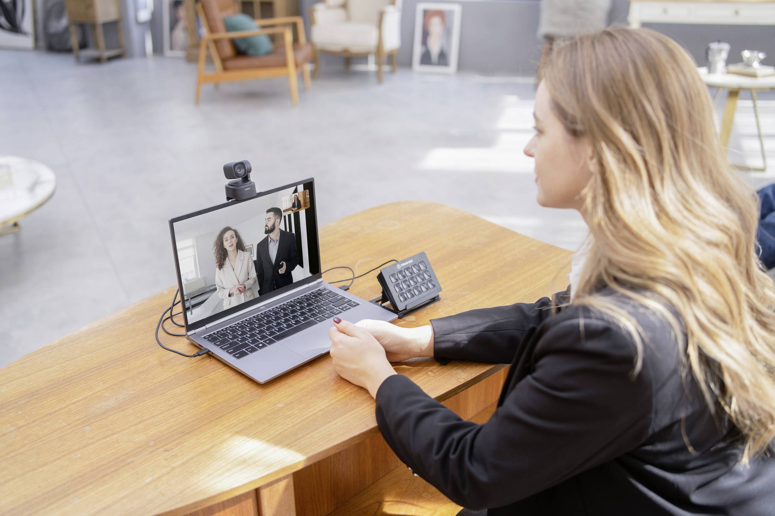 A woman is sitting at a table and participating in a video conference call on a laptop. Two people are visible on the screen.