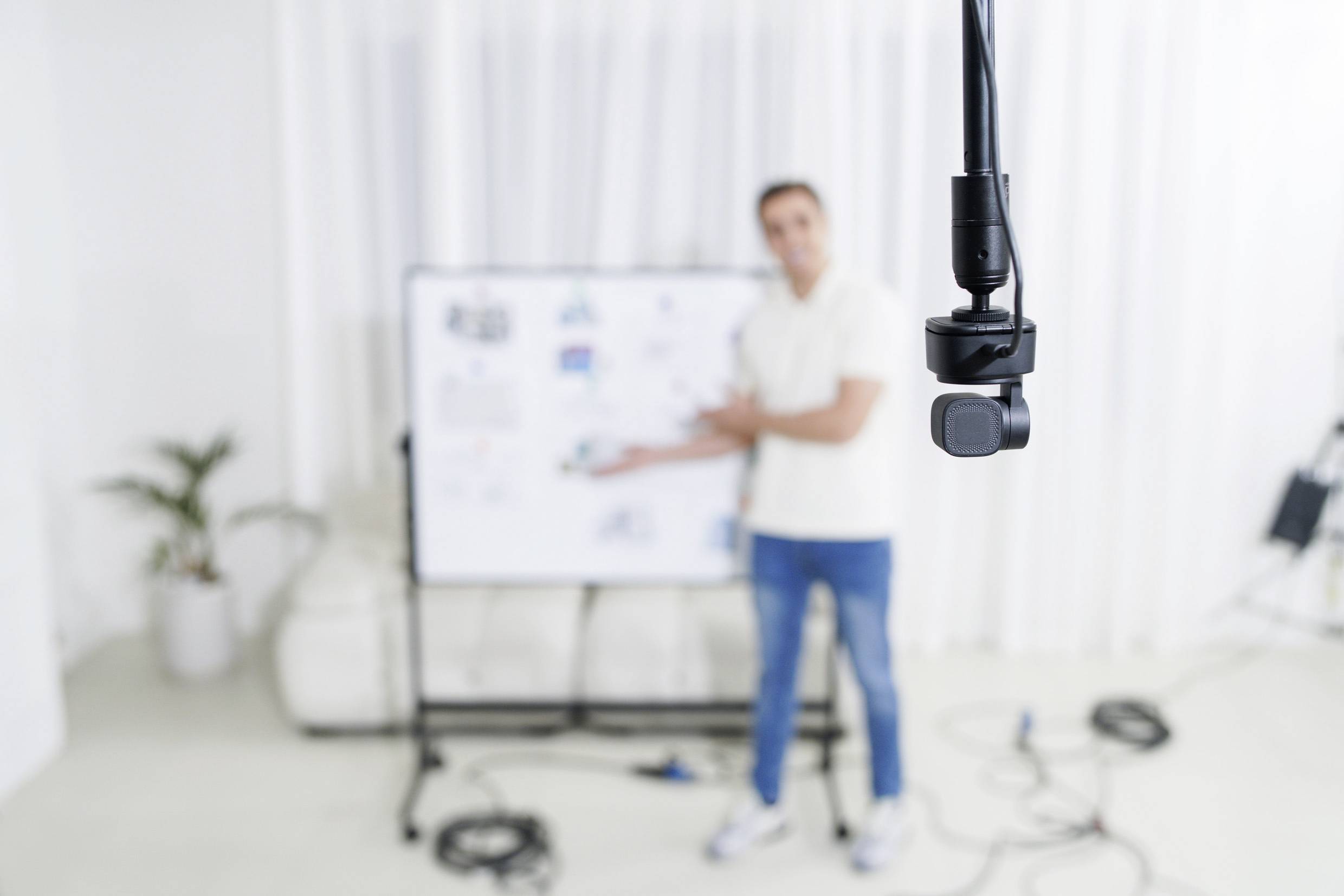 Man in the background pointing at a whiteboard; a microphone in the foreground. The environment is a bright room with cables on the floor.
