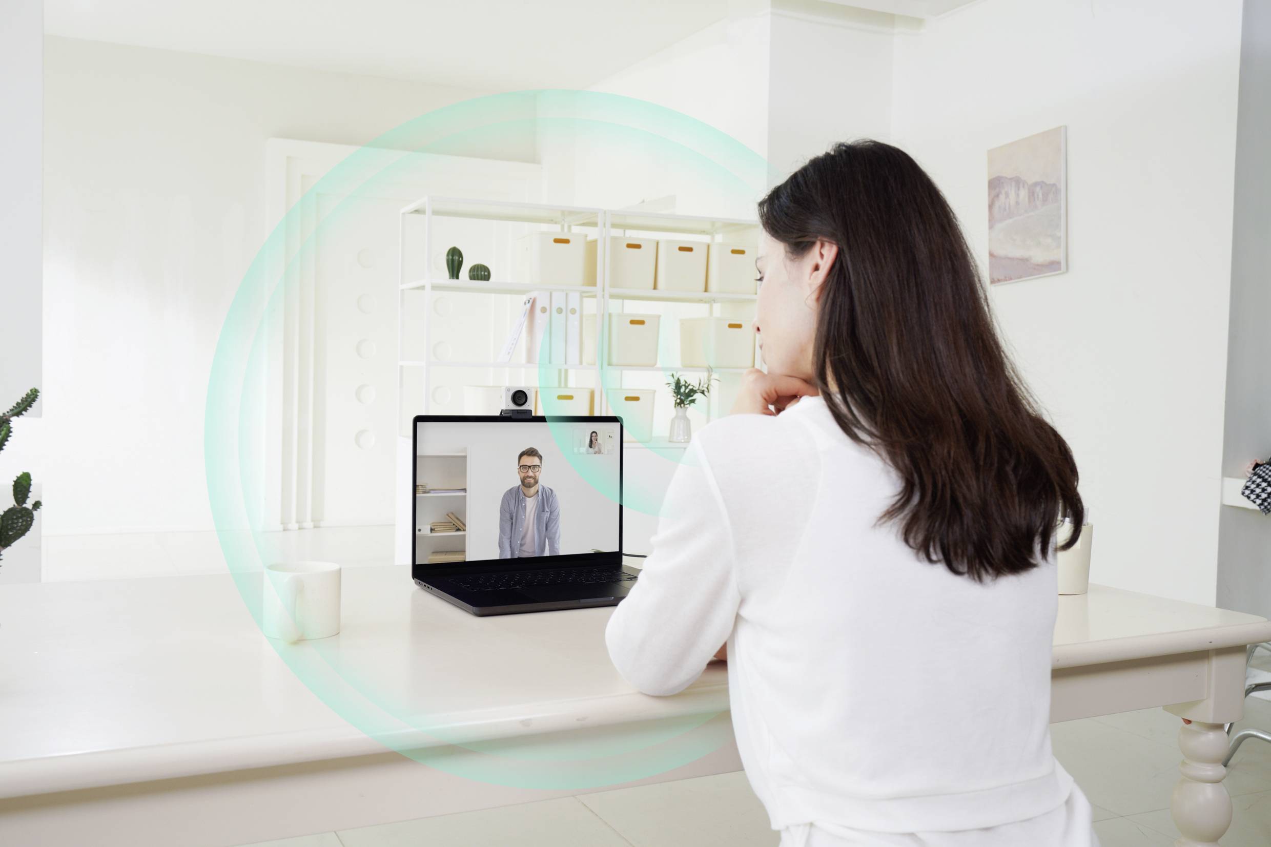 Woman in white room during video call with a person on a laptop; bookshelf in the background.