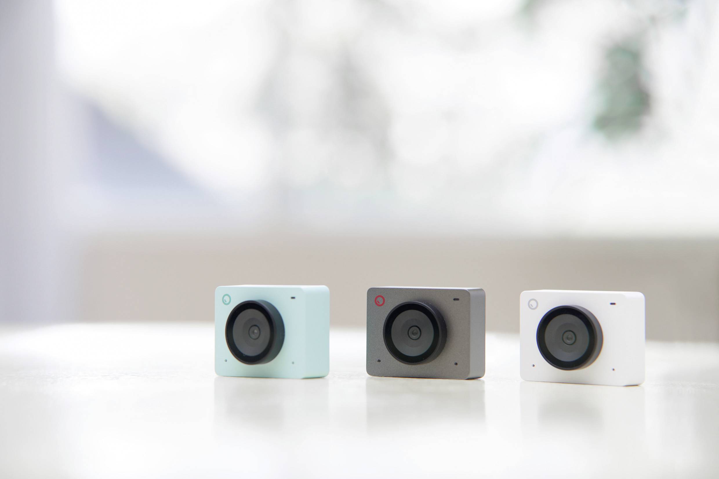 Three square cameras in pastel colours sit side by side on a table, against a blurred background.