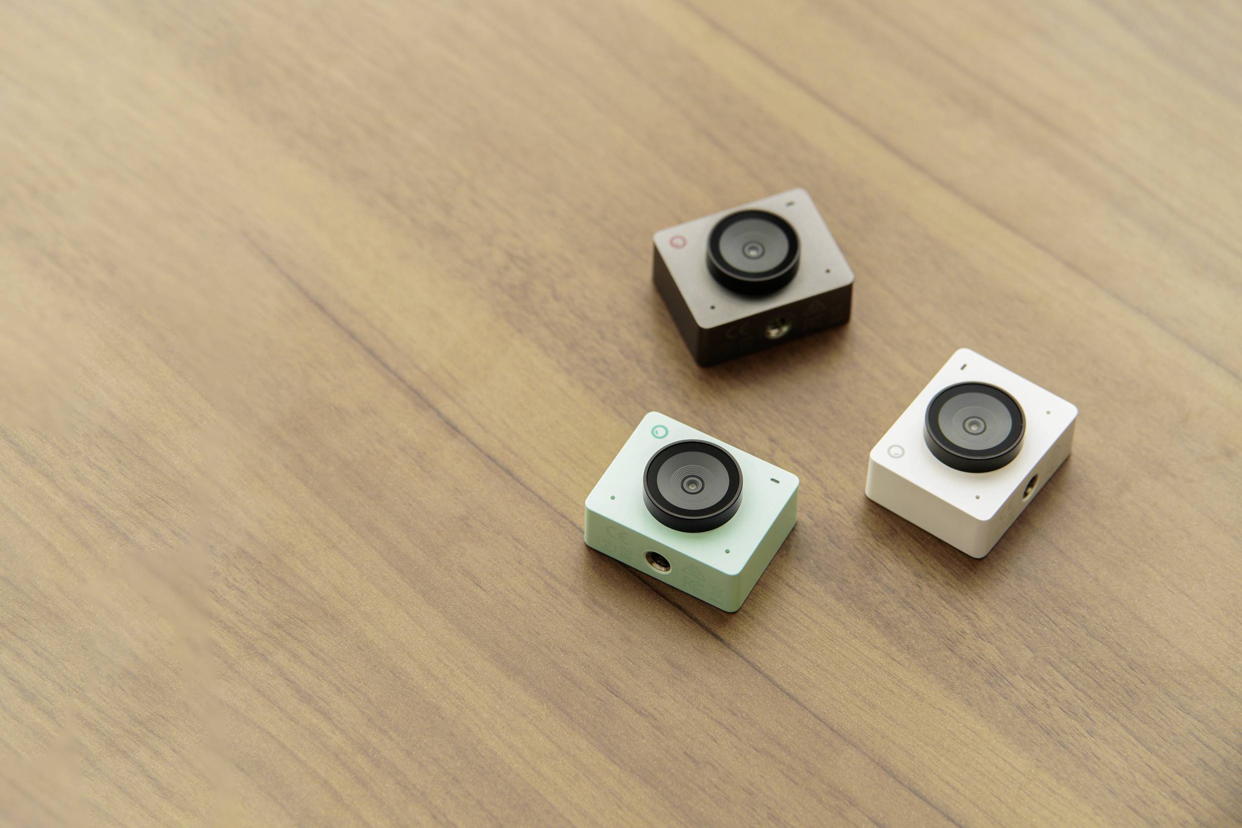 Three small action cameras in different colours (black, mint green, white) are lying on a wooden table.