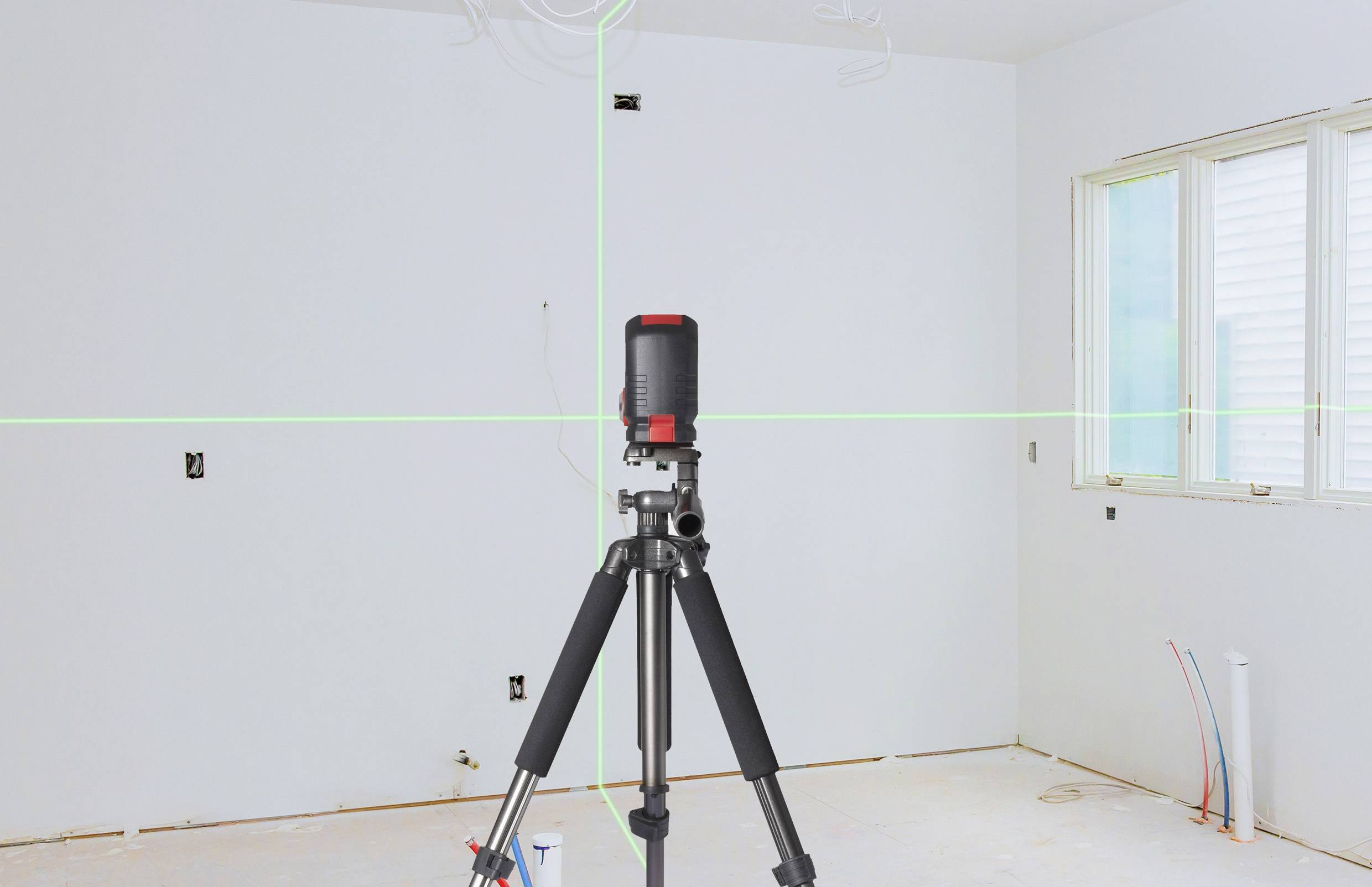 A laser measuring device stands on a tripod in an empty room. Green laser lines project reference lines onto the walls and ceiling.