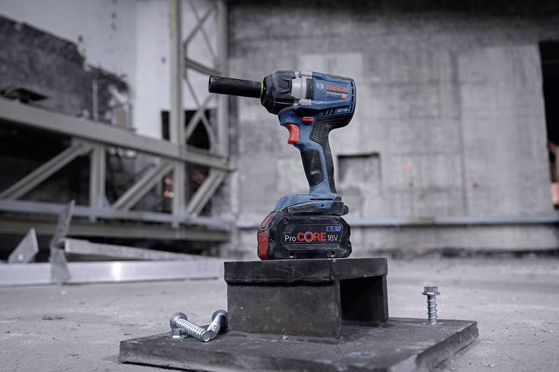 Cordless drill sits on a building site, surrounded by screws and metal parts. In the background are concrete walls and steel beams.