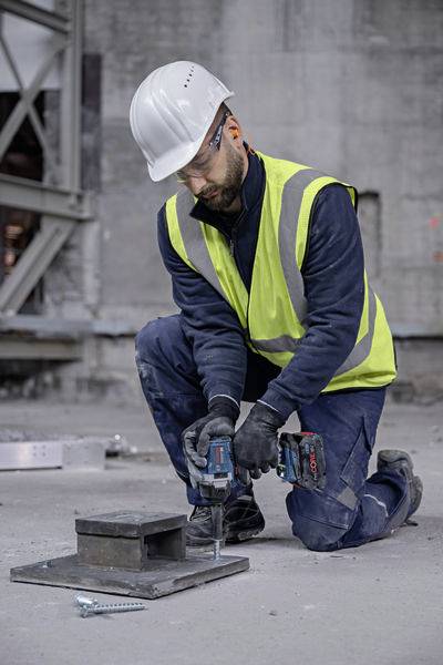 A construction worker wearing a hard hat and high-visibility vest is kneeling on a concrete floor and using an electric drill to secure metal plates.