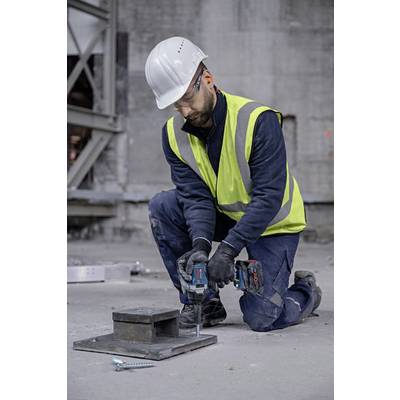 A construction worker wearing a hard hat and high-visibility vest is kneeling on a concrete floor and using an electric drill to secure metal plates.