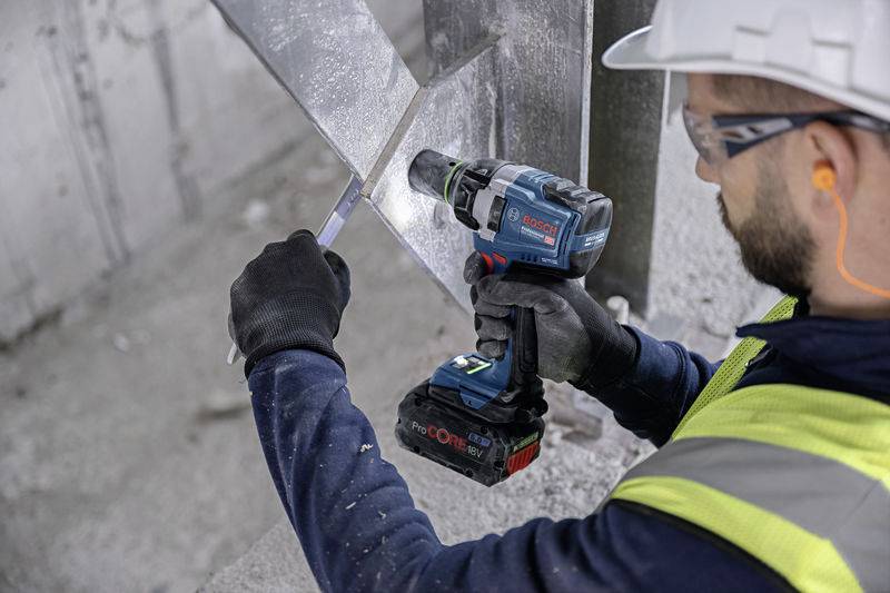 A construction worker wearing a high-visibility safety vest is using an electric drill to work on metal. He is wearing a hard hat, gloves, and ear protection.