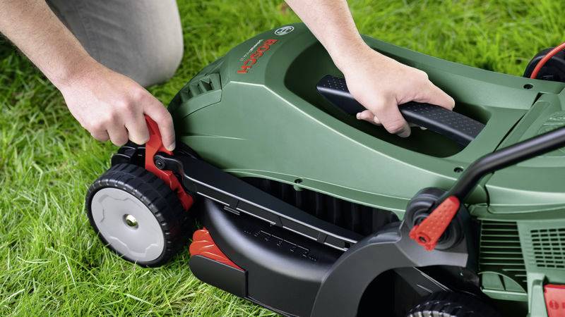 A person is adjusting the cutting height on a green electric lawnmower on a lawn area.