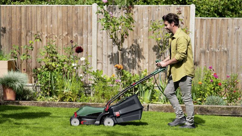 A man in a garden is mowing the lawn with a green lawnmower. Colourful flowers and a wooden fence are in the background.