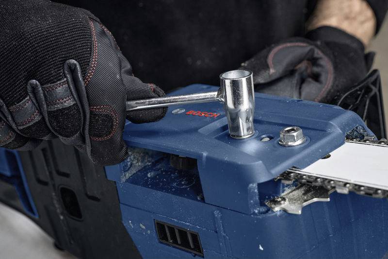 A person wearing black gloves is turning a screw on a blue chainsaw with a spanner.