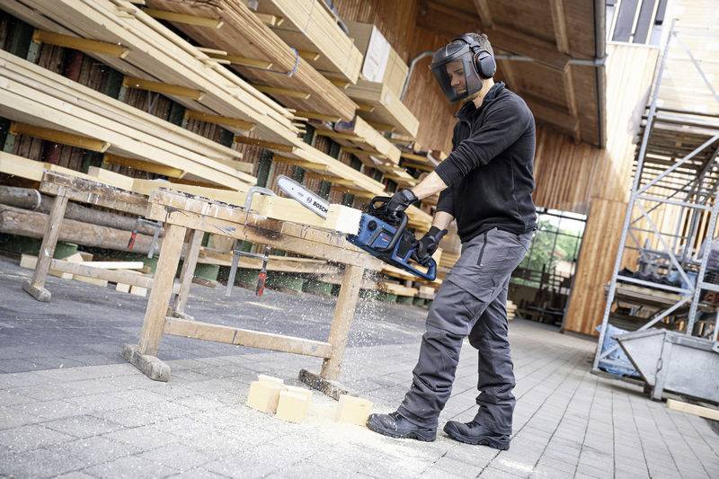A person wearing protective equipment is cutting wood on a sawborse with a chainsaw. Stacked timber is visible in the background.