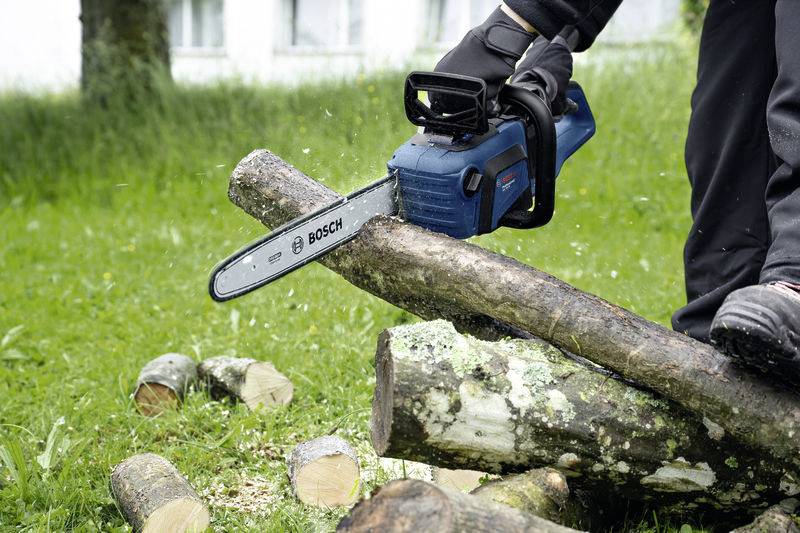 A person is sawing a wooden log outside with a blue chainsaw. Grass and cut pieces of wood are visible in the background.