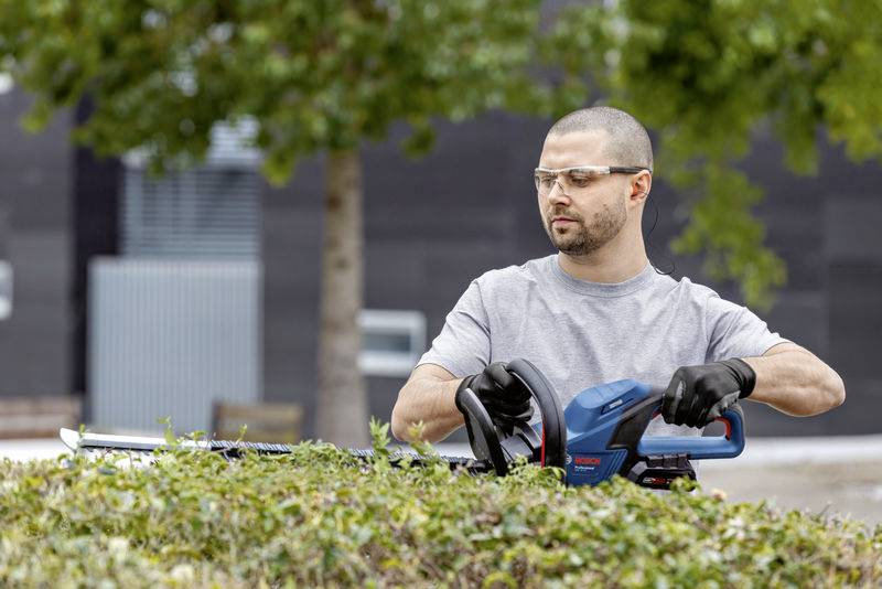 A man wearing safety glasses and gloves is trimming a hedge with an electric hedge trimmer in a garden.