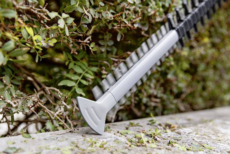 A hedge trimmer is cutting a green bush. Leaves and branches are visible in the foreground. The scene depicts gardening work.