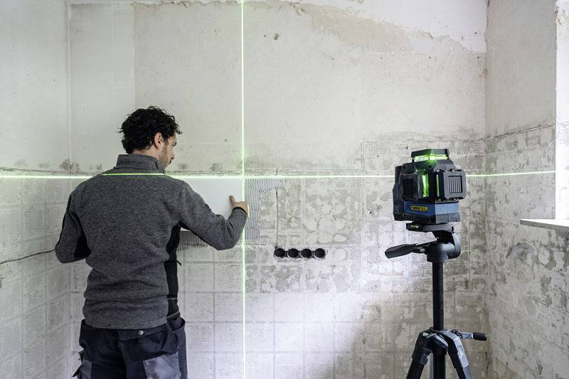 A man is aligning tiles using a cross-line laser on the wall of a room that needs renovation.