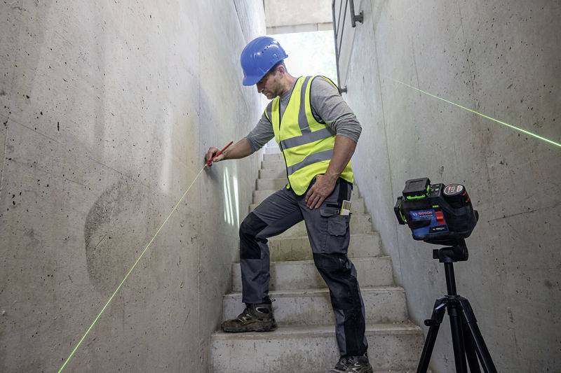 A construction worker wearing a blue hard hat and yellow high-visibility vest is marking a wall precisely with a laser device in a stairwell.