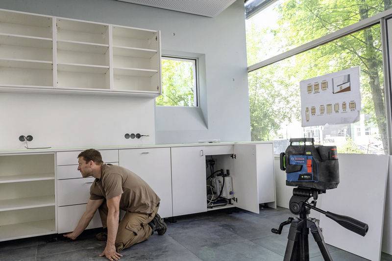 A man is installing cupboards in a modern kitchen. In the foreground, a measuring device stands on a tripod.