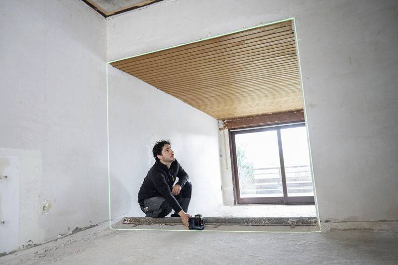 A person is using a laser measuring device in an unfinished room to align the walls. The room has bare walls and a wooden floor.