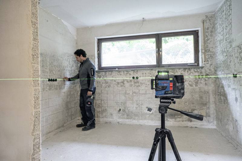 A tradesman measures the wall with a laser in an empty room with tiled walls and windows. A laser device is mounted on a tripod in the foreground.