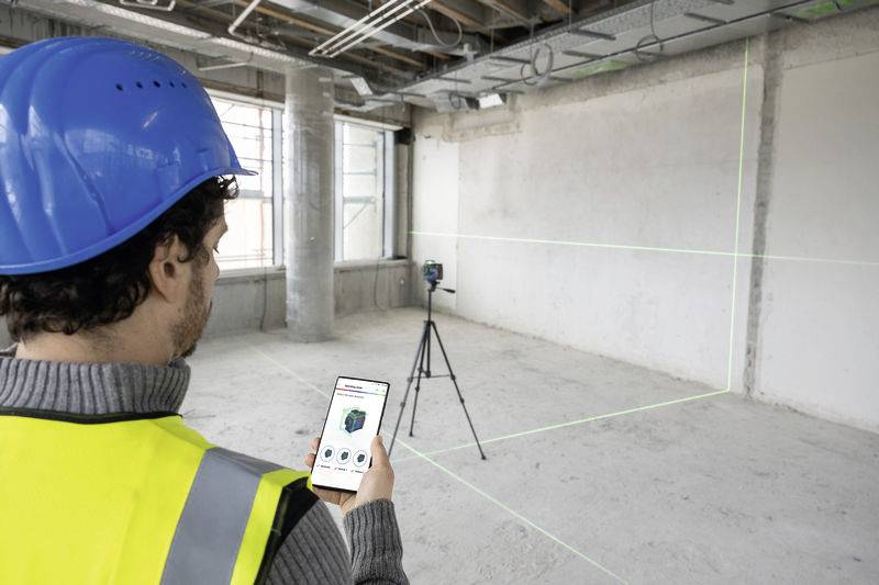 A construction worker wearing a hard hat and high-visibility vest is using a smartphone to control a surveying device mounted on a tripod in an empty room.