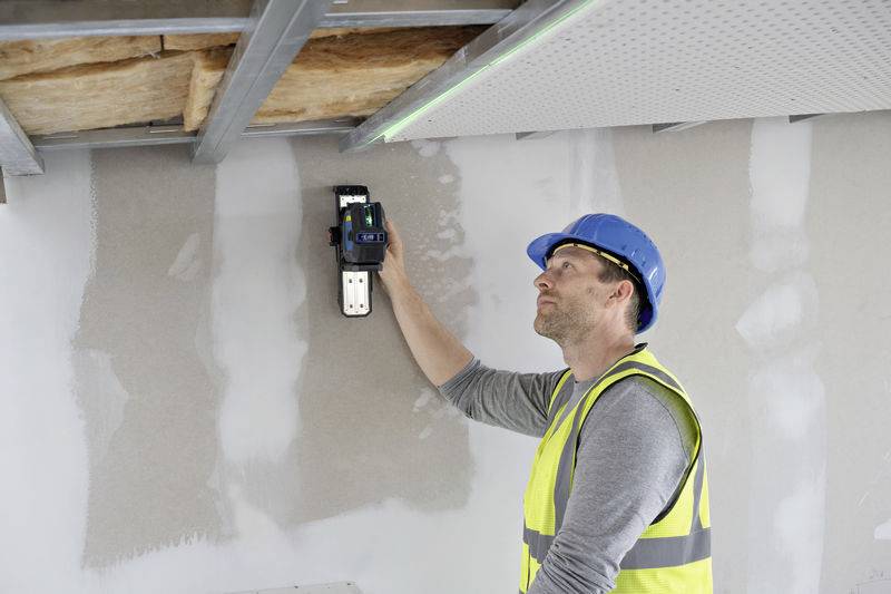 A construction worker in a high-visibility vest and hard hat is using a digital measuring device on a wall to take measurements.