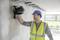 A construction worker is wearing a blue hard hat and a yellow high-visibility vest. He is working with a laser tool on a white wall.