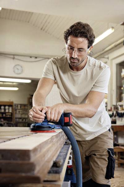 A man is sanding a wooden board in a workshop using an electric sanding tool. Shelves are visible in the background.