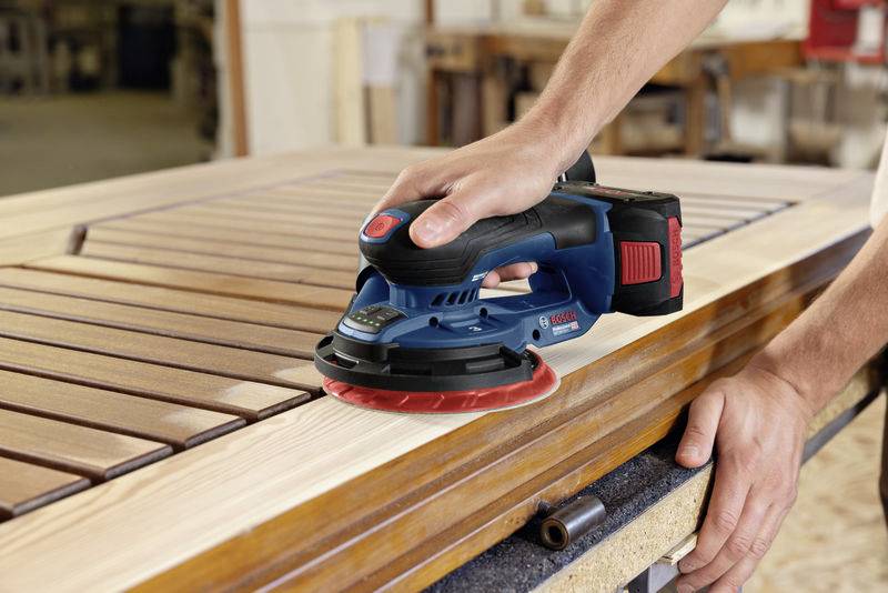 A person is sanding the wooden surface of a piece of furniture with an electric sanding tool in a workshop.