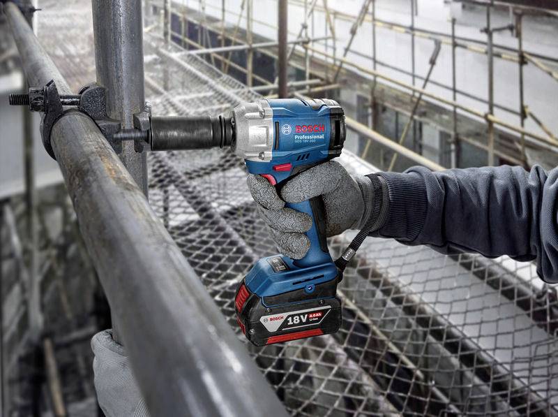 A person is using a blue cordless impact driver to secure a screw on a metallic scaffolding. In the background, the scaffolding of a construction site is visible.