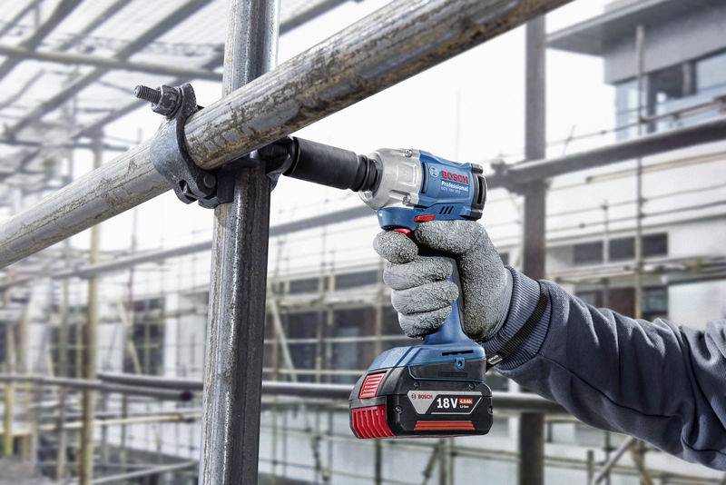 A person is working with a blue, battery-powered screwdriver on scaffolding outdoors. A building is visible in the background.