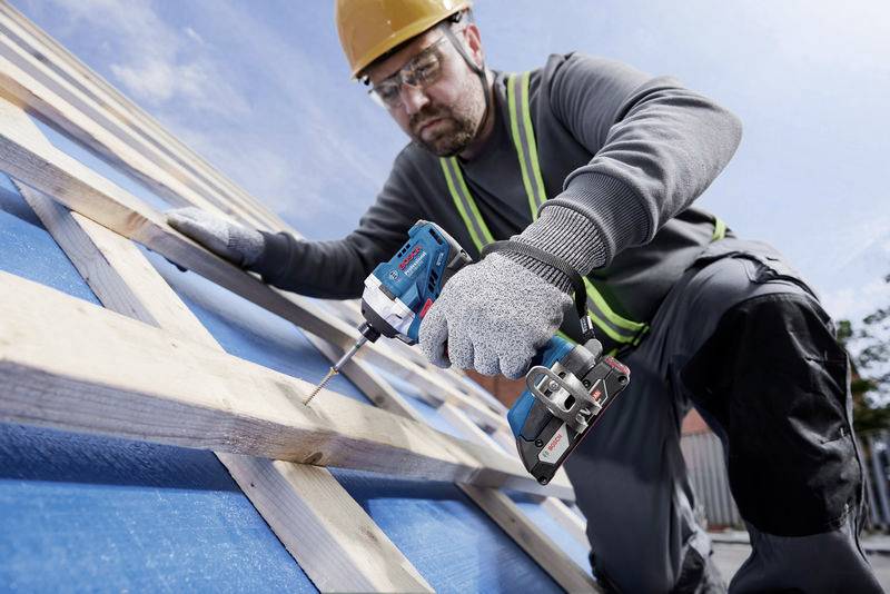 A construction worker wearing a hard hat and gloves is using a cordless drill to secure wooden boards on a sloping roof.