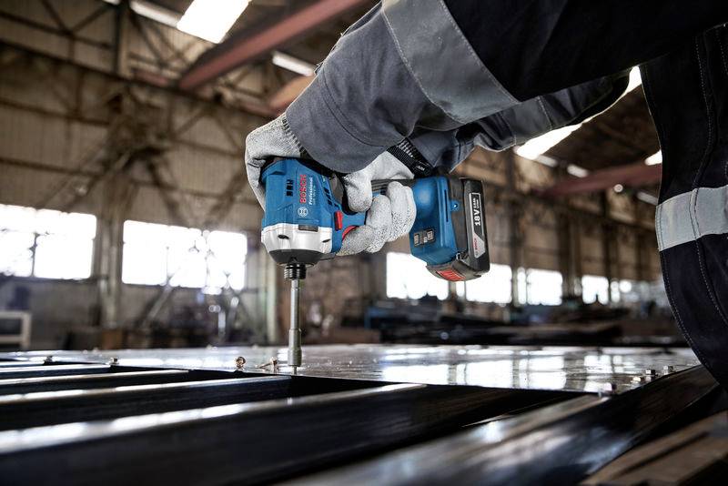 A person in a workshop is using a blue cordless drill to drive screws into a metal plate. The background shows an industrial warehouse.