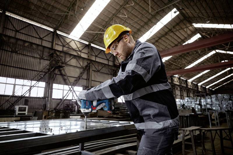 A construction worker wearing a hard hat is drilling into a metal structure using a power drill in a large, industrial warehouse.