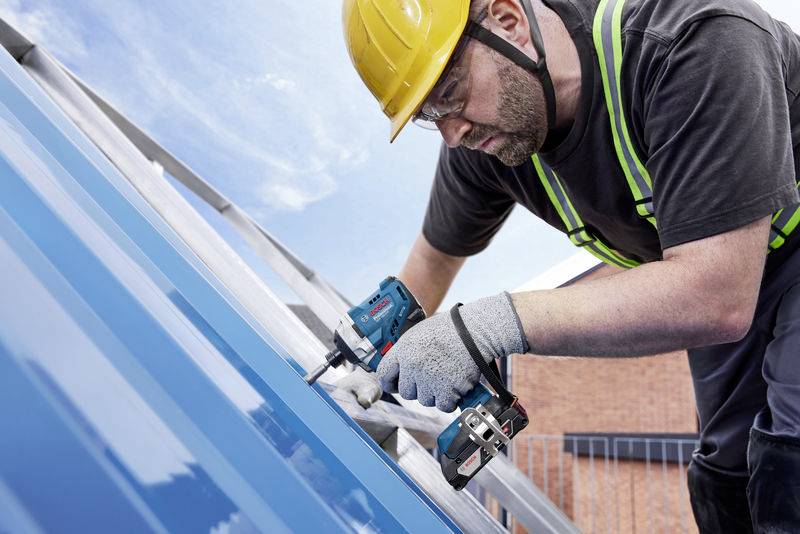 A construction worker wearing a hard hat and high-visibility vest is working with an electric power tool on a blue metal roof outdoors.