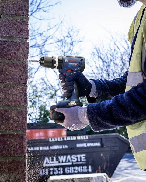 A person is drilling a hole in a brick wall using an electric drill. A skip is visible in the background.