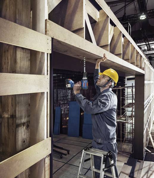 A construction worker in protective gear is drilling a hole in a wooden scaffolding on a building site using an electric drill.