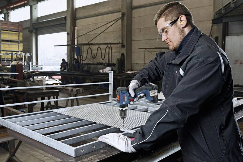 A man is working in a workshop, drilling a metal grid on an industrial workbench. He is wearing protective clothing and safety glasses.
