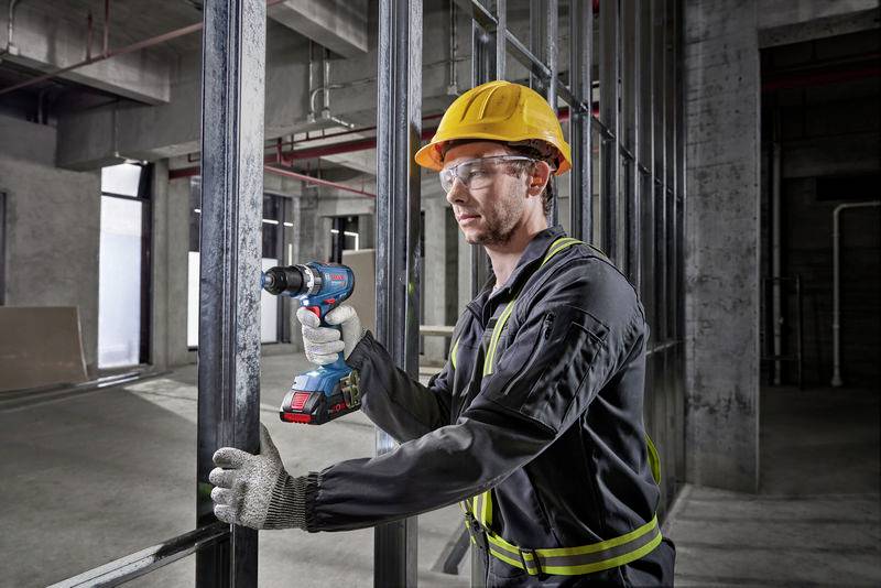 A construction worker wearing a yellow hard hat and safety glasses is using a power drill to secure steel beams in a construction site environment.