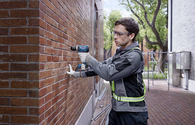 A man wearing safety glasses and gloves is drilling into a red brick wall with a blue drill. Trees are visible in the background.