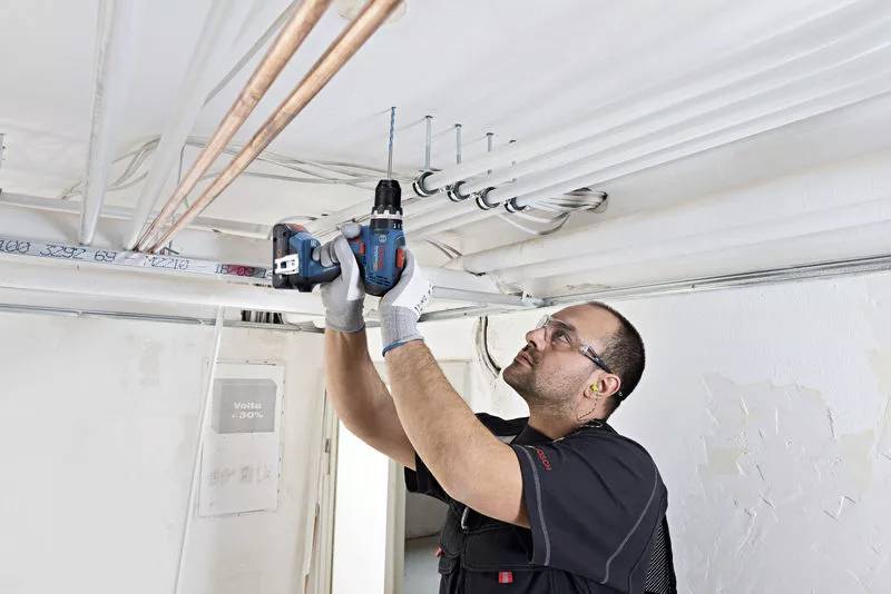 A worker fixing several cables and pipes to a ceiling using a drill. He is wearing protective clothing and gloves.