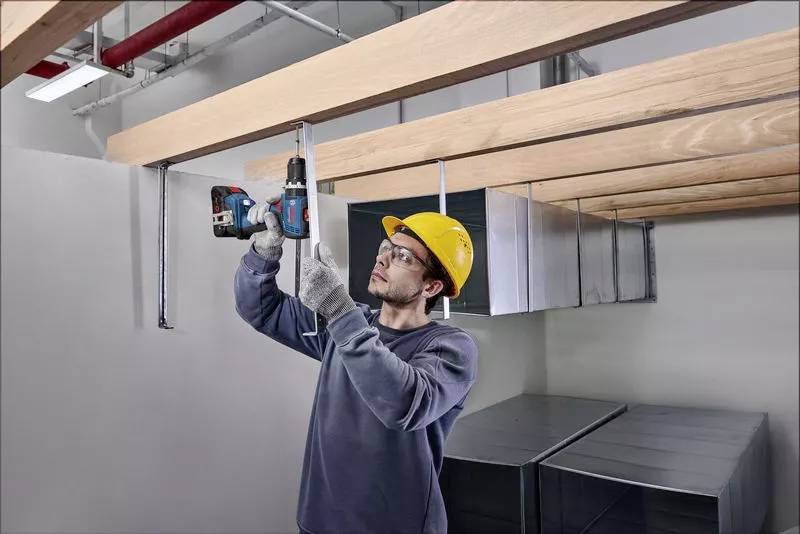 A worker wearing a hard hat is securing wooden beams to the ceiling using a drill in an industrial interior.