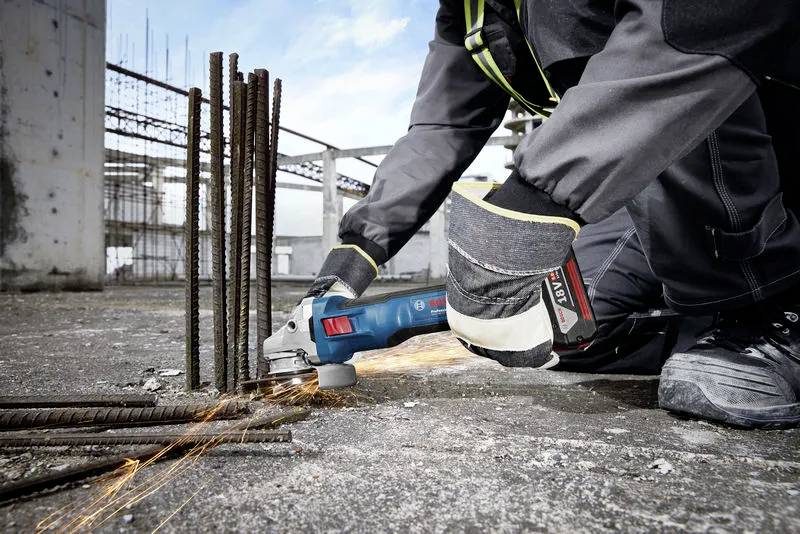 A worker in protective gear cuts metal rods on a construction site using an angle grinder, with sparks flying.