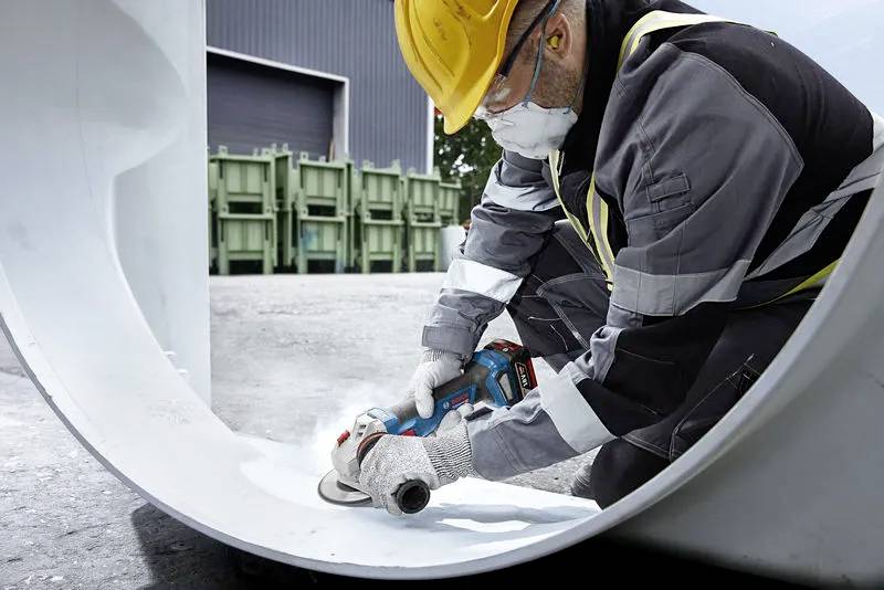 A worker wearing a yellow hard hat and protective gear uses a power tool to grind the inside of a large white pipe in an industrial area.